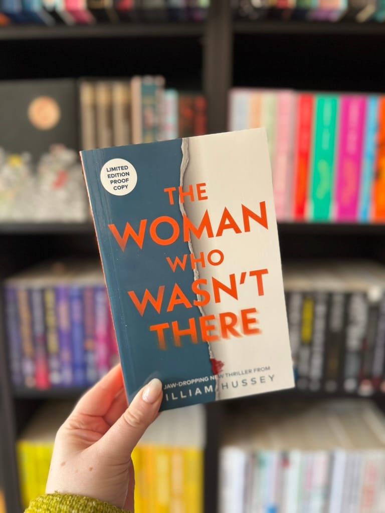 A hand holds a book titled "The Woman Who Wasn't There" by William Hussey. Background shows colorful bookshelves, creating a vibrant scene.
