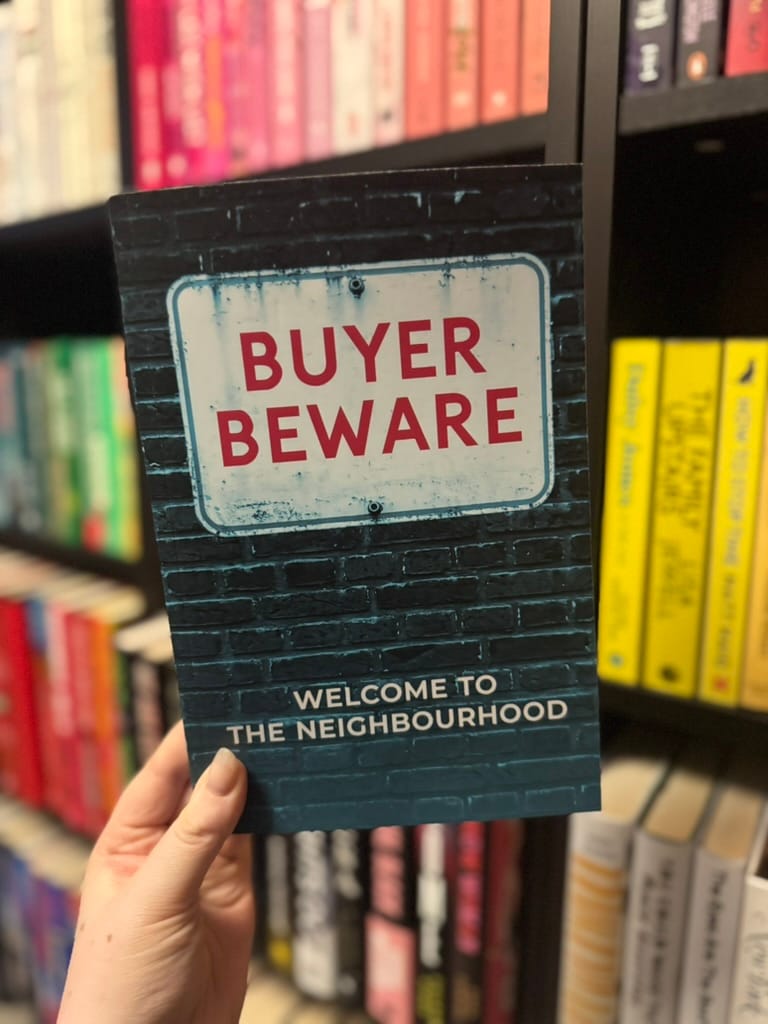 A hand holds a book titled "Buyer Beware: Welcome to the Neighbourhood" against a bookshelf with colorful spines.