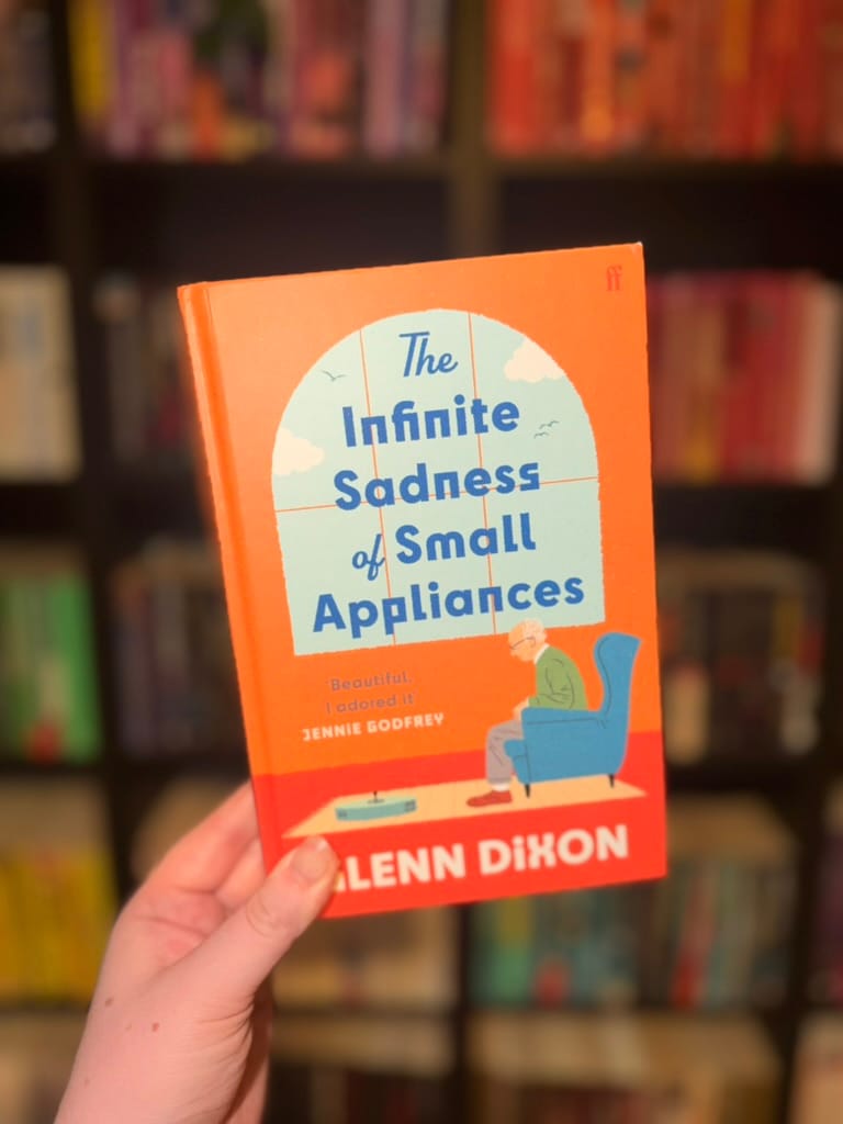 A hand holds a copy of The Infinite Sadness of Small Appliances by Alann Dixon in front of softly blurred rainbow bookshelves.