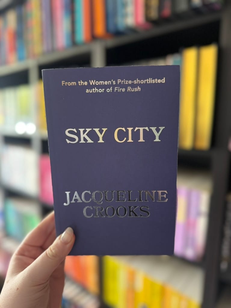 A hand holds the proof copy of Sky City by Jacqueline Crooks in front of softly blurred rainbow bookshelves. T