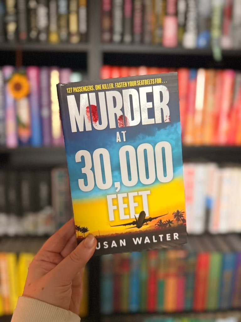 Hand holding the book Murder at 30,000 Feet by Susan Walter in front of colourful blurred bookshelves.