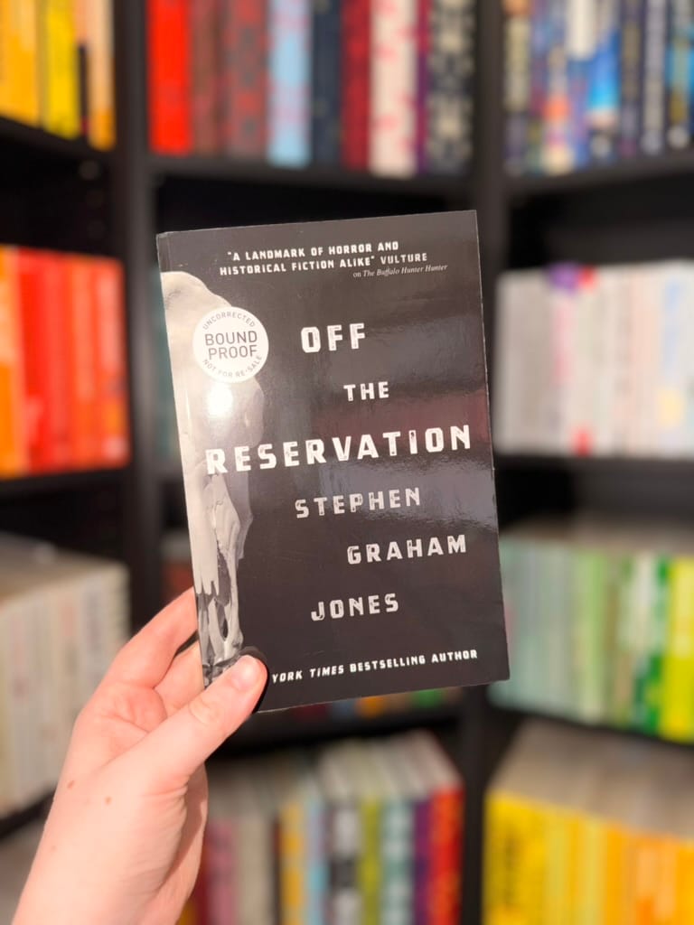 A hand holds a bound proof copy of Off the Reservation by Stephen Graham Jones in front of softly blurred, colour-organised bookshelves. 