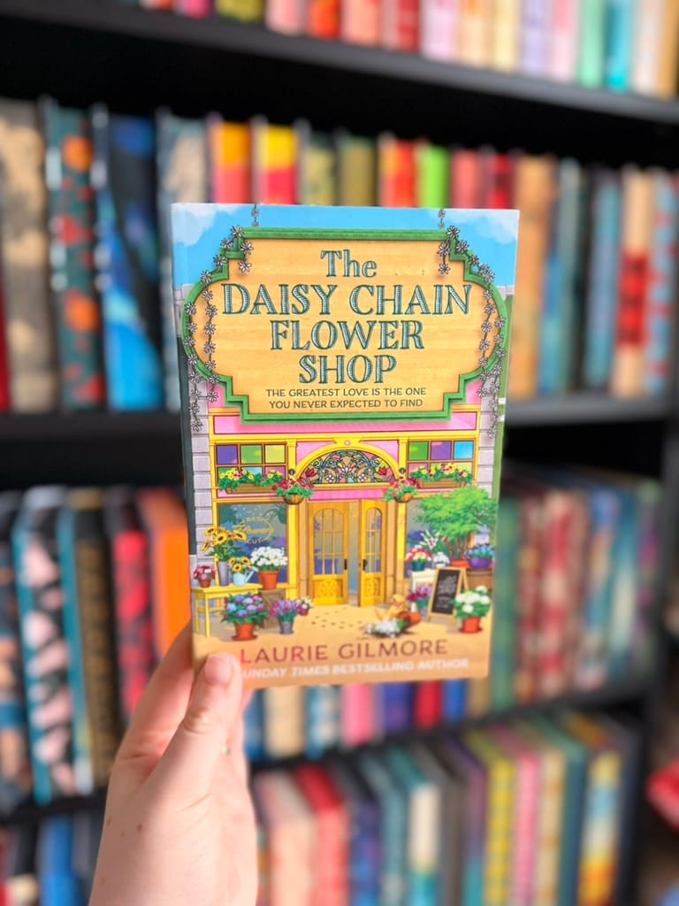 A hand holds The Daisy Chain Flower Shop by Laurie Gilmore in front of softly blurred rainbow bookshelves. 