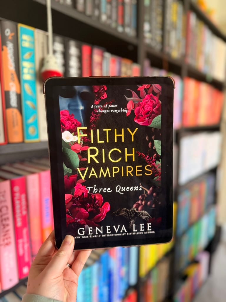 A hand holds a tablet displaying the cover of Three Queens from Geneva Lee’s Filthy Rich Vampires series.