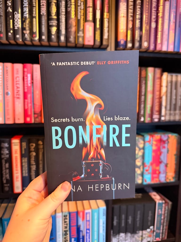 A hand holds a copy of Bonfire by Anna Hepburn in front of colour-coordinated bookshelves.