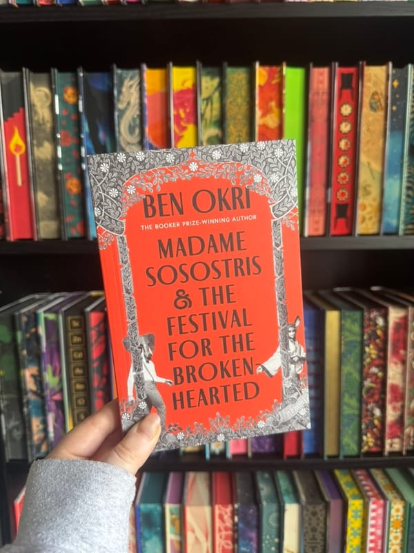 A hand holds a proof copy of Madame Sosostris & The Festival for the Brokenhearted by Ben Okri in front of softly blurred rainbow bookshelves.