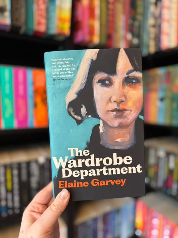 A hand holds a copy of The Wardrobe Department by Elaine Garvey in front of softly blurred rainbow bookshelves.