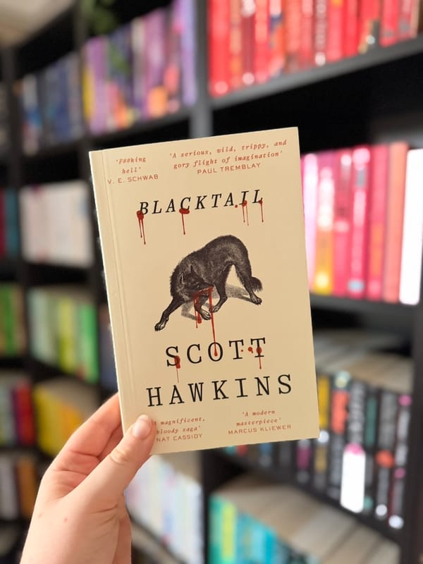 Blacktail by Scott Hawkins in front of softly blurred rainbow bookshelves. 