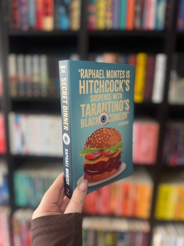 A hand holds a book titled "Secret Dinner" with a burger illustration. Bookshelves in the blurred background create a colorful setting.