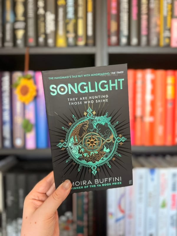A hand holds a copy of Songlight by Moira Buffini in front of softly blurred rainbow bookshelves. 