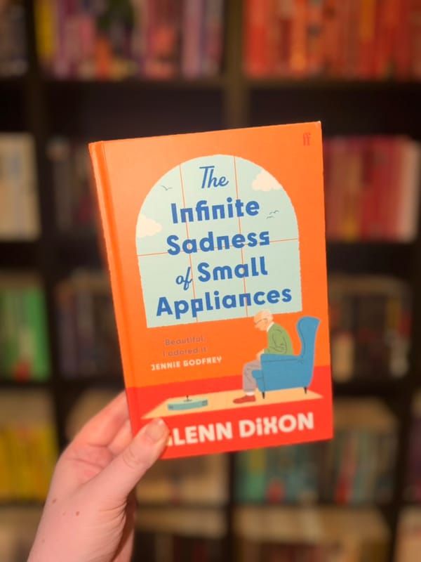 A hand holds a copy of The Infinite Sadness of Small Appliances by Alann Dixon in front of softly blurred rainbow bookshelves.