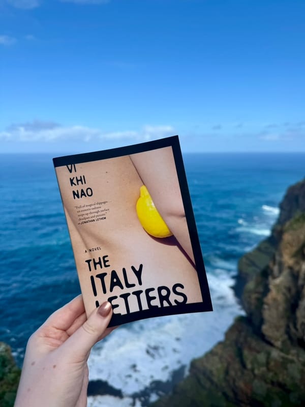 Hand holding "The Italy Letters" book against a clear blue sky and ocean cliffside background. Cover shows a lemon on skin.