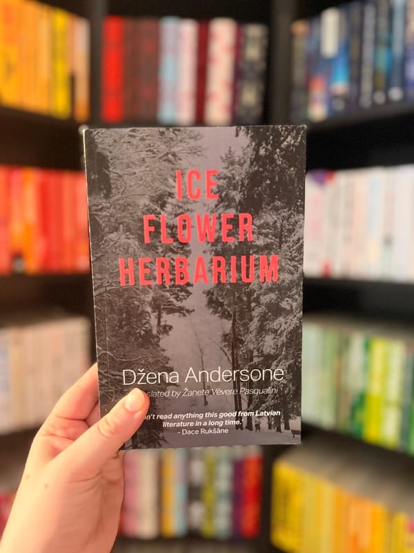 A hand holds a copy of Ice Flower Herbarium by Džena Andersone in front of a softly blurred bookshelf filled with colour-coordinated books. 
