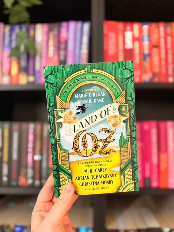 A hand holds Land of Oz, a green and gold anthology edited by Marie O’Regan and Paul Kane, in front of blurred, colour-coordinated bookshelves.