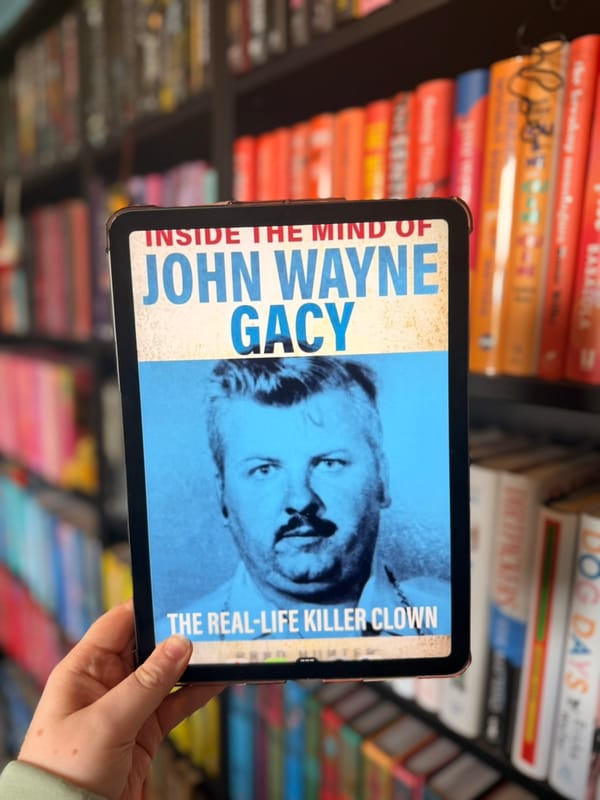 A hand holds an e-reader displaying the cover of Inside the Mind of John Wayne Gacy: The Real-Life Killer Clown by Christopher Berry-Dee in front of softly blurred rainbow bookshelves. 