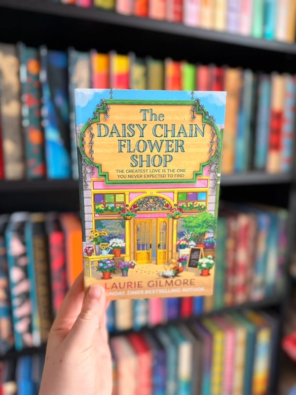 A hand holds The Daisy Chain Flower Shop by Laurie Gilmore in front of softly blurred rainbow bookshelves. 