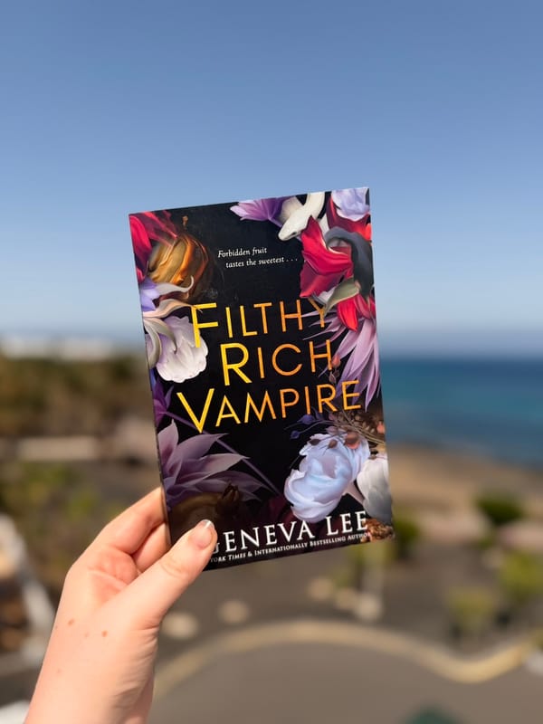 A hand holds a copy of Filthy Rich Vampire by Geneva Lee outdoors against a softly blurred coastal backdrop. 