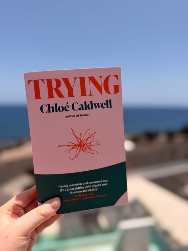 A hand holds a copy of Trying by Chloé Caldwell outdoors against a softly blurred coastal backdrop.