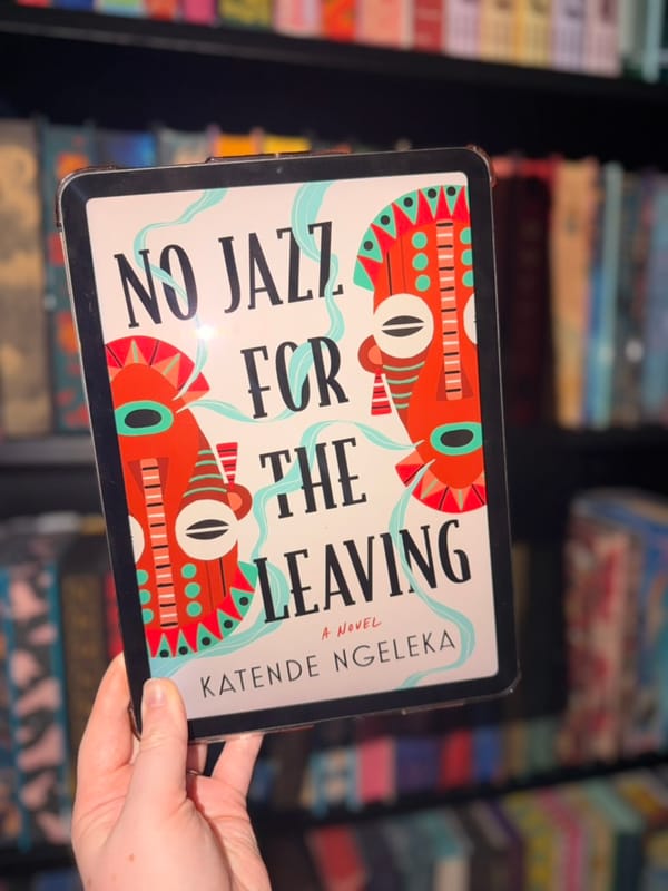 A hand holds an e-reader displaying the cover of No Jazz for the Leaving by Katende Ngeleka in front of softly blurred rainbow bookshelves. 
