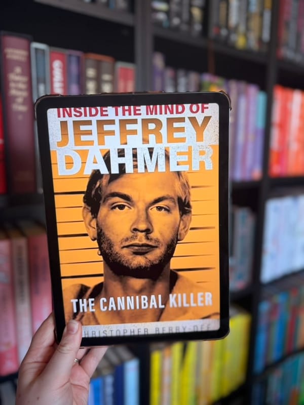 A hand holds an e-reader displaying the cover of Inside the Mind of Jeffrey Dahmer: The Cannibal Killer by Christopher Berry-Dee in front of softly blurred rainbow bookshelves.
