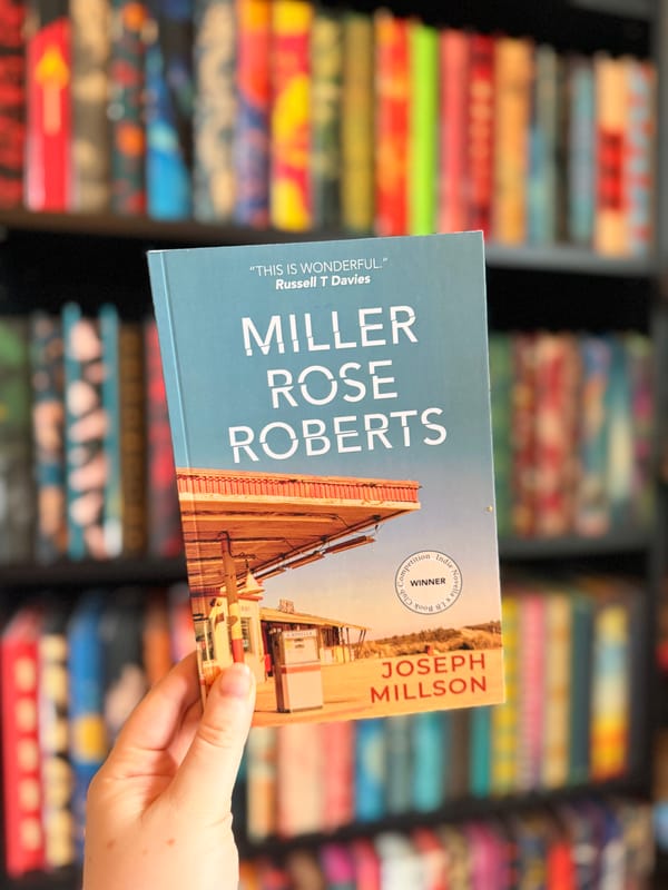 A hand holding a copy of Miller Rose Roberts by Joseph Millson in front of a colourful, softly blurred bookshelf filled with books.