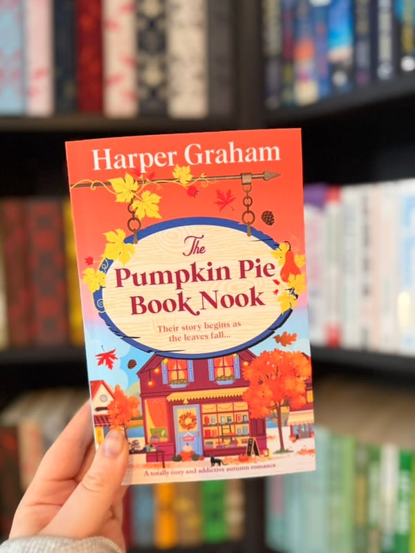 A hand holds a copy of The Pumpkin Pie Book Nook by Harper Graham in front of blurred bookshelves. 