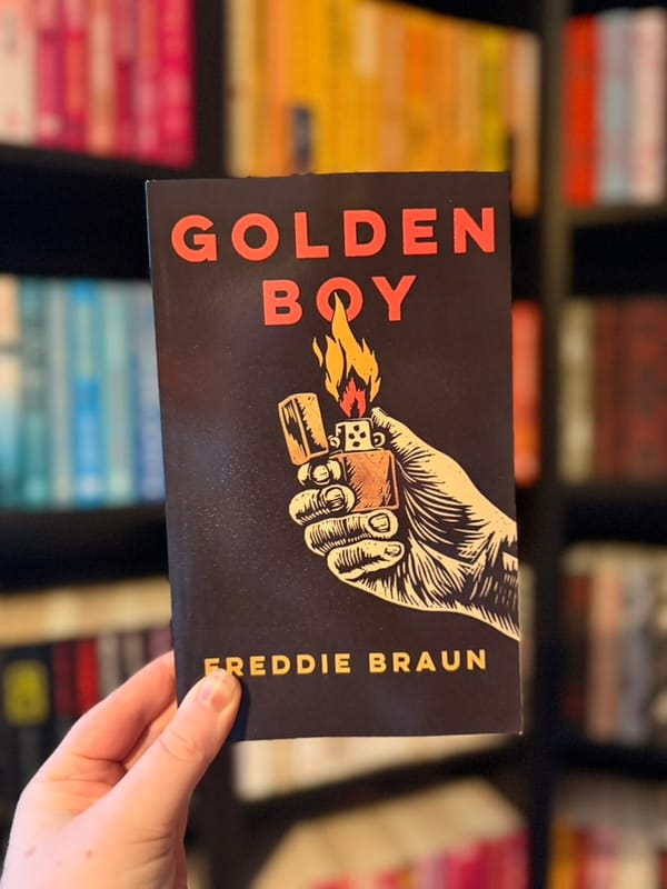 A hand holds a copy of Golden Boy by Freddie Braun in front of colour-coordinated bookshelves.