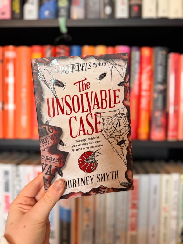 A hand holds a copy of The Unsolvable Case by Courtney Smyth in front of blurred bookshelves.