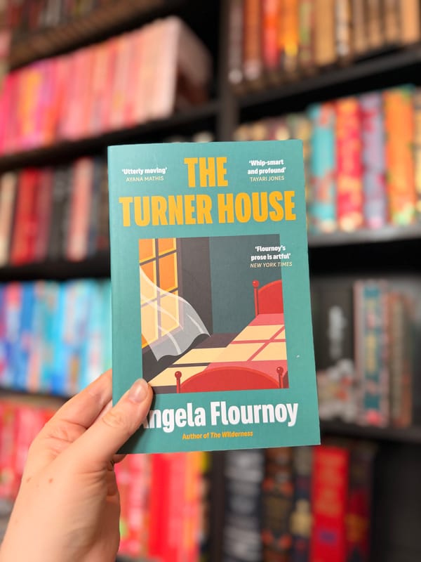 A hand holding a copy of The Turner House by Angela Flournoy in front of colourful, blurred bookshelves. 