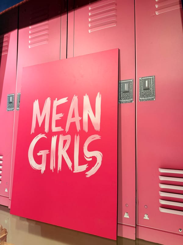 Pink school lockers with a large hot-pink Mean Girls sign featuring the title in white brush lettering.